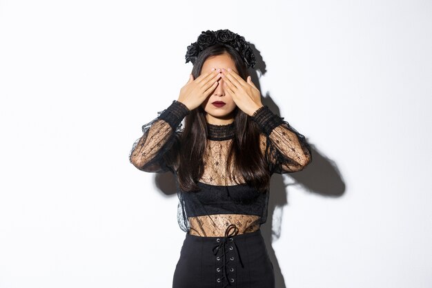 Free photo image of woman wearing gothic wreath and black lace dress shut her eyes with hands, waiting for surprise on halloween, standing over white background with patient expression.