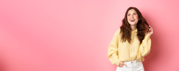 Image of young 20s woman laughing and playing with her hair looking aside at upper left corner promotion standing happy against pink background