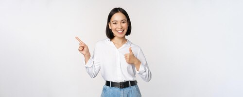 Image of young asian business woman smiling while pointing finger left and showing thumbs up recommending product praise standing over white background