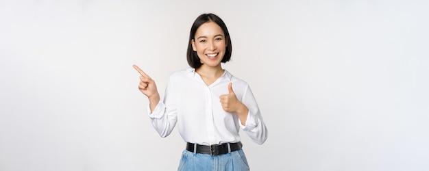 Image of young asian business woman smiling while pointing finger left and showing thumbs up recommending product praise standing over white background