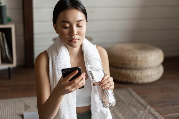 Image of young asian fitness girl with fit body towel wrapped around neck looking at mobile phone ap...