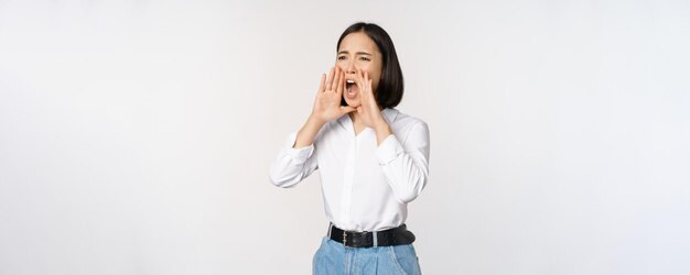 Image of young asian woman calling for someone shouting loud and searching around standing against w