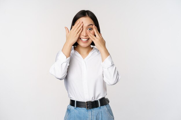 Image of young asian woman close her eyes and smiles waits for surprise anticipates peeking through fingers stands over white background