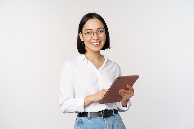 Изображение молодой азиатской работницы компании в очках, улыбающейся и держащей цифровой планшет, стоящий на белом фоне