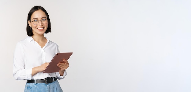 Image of young asian woman company worker in glasses smiling and holding digital tablet standing over white background