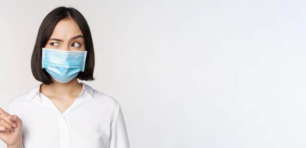 Изображение молодой азиатки в медицинской маске, указывающей влево и смотрящей с подозрительным смущенным выражением лица, стоящей на белом фоне