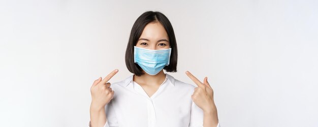 Image of young asian woman pointing at herself while wearing medical face mask concept of covid19 protection standing over white background