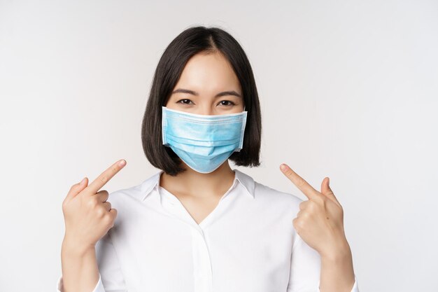 白い背景の上に立っているcovid19保護の医療フェイスマスクの概念を身に着けている間自分自身を指している若いアジアの女性の画像