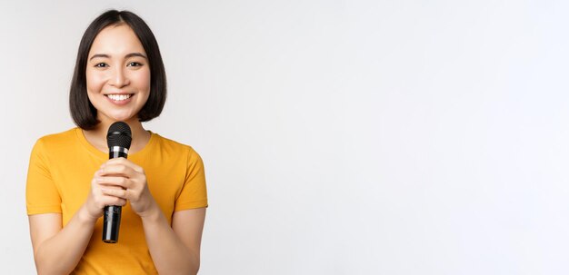 Image of young asian woman talking in microphone perfom with mic giving speech standing in yellow tshirt against white background