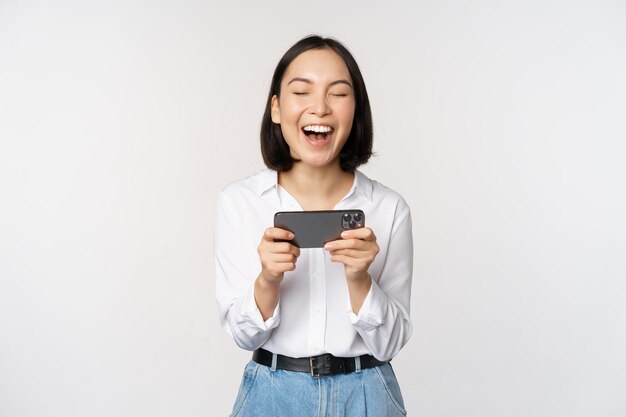 스마트폰 앱에서 휴대폰을 들고 화면을 바라보며 흰색 배경 위에 서서 웃고 웃고 있는 젊은 아시아 여성의 이미지
