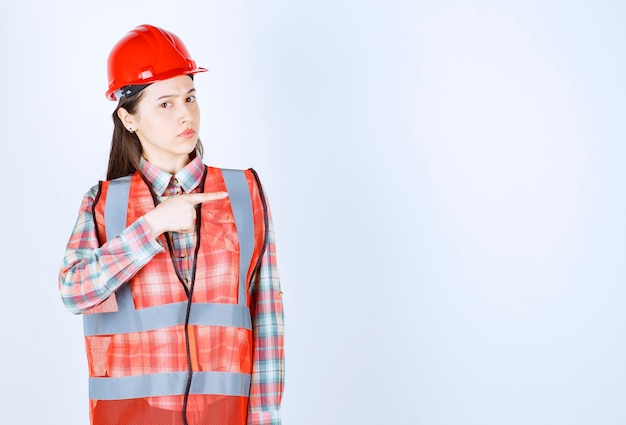 Image of young beautiful female engineer in helmet pointing at somewhere. 