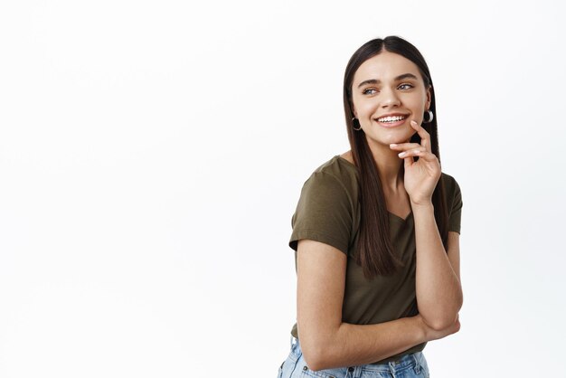 Immagine di giovane bella modella femminile con lunghi capelli scuri che guarda a sinistra e sorride con gioia fissando lo spazio della copia del logo toccando il viso naturale con trucco leggero in piedi su sfondo bianco