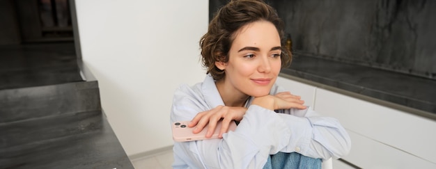 Immagine di una giovane donna brunetta seduta sognante in cucina sorridendo pensierosa con in mano uno smartphone