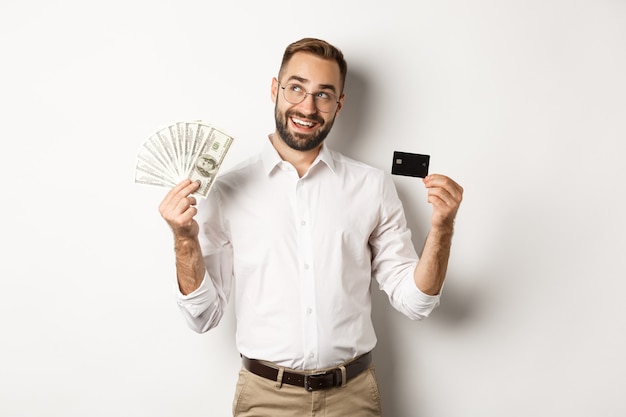 Image of young businessman holding credit card and money, looking at upper left corner and thinking about shopping, standing  