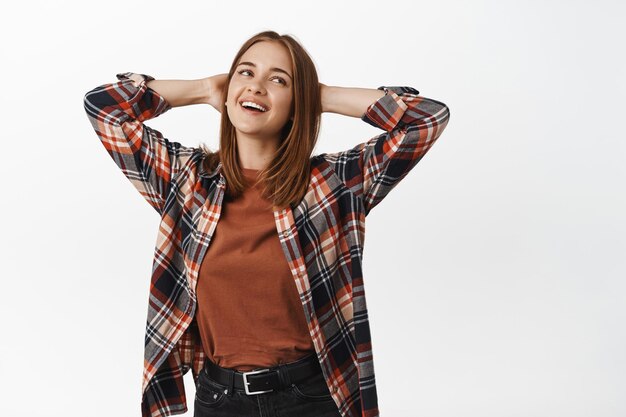 Image of young carefree woman, enjoying free time, leisure and vacation time, hold hands behind head, smiling with satisfied and relaxed face, standing over white background