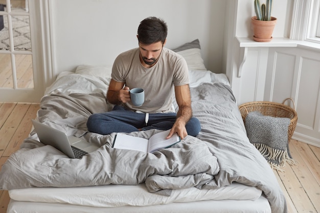 Image of young Caucasian man has morning coffee, sits crossed legs on bed