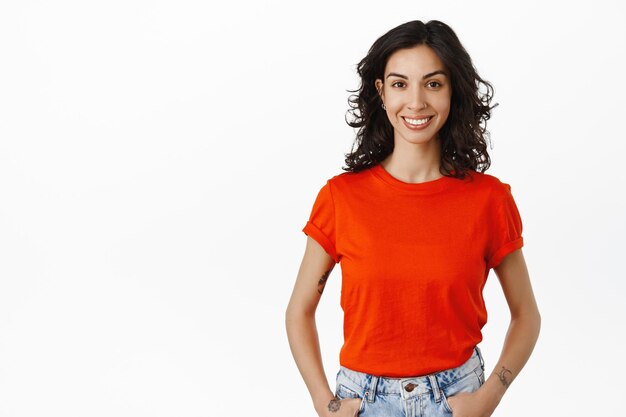 Image of young caucasian woman with curly dark hair, stands in red t-shirt with happy relaxed smile, hold hands in jeans, isolated over white background.