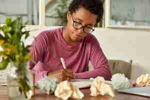 Free photo image of young dark skinned teacher prepares for conducting lesson at college, makes notes in notepad, surrounded with paper balls, has creative chaos on table, focused down, wears optical glasses