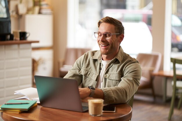 안경을 쓴 젊은 디지털 유목민 남자가 카페에서 앉아 커피에서 노트북을 사용합니다.