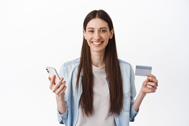 Image of young female model holding credit card and smartphone, concept of online shopping, contactless payment and internet delivery, standing over white wall