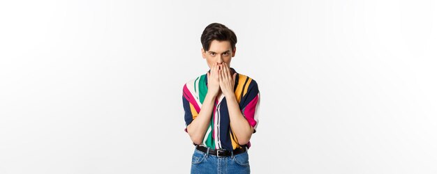 Image of young gay man looking intrigued holding hands on mouth gossiping standing over white background