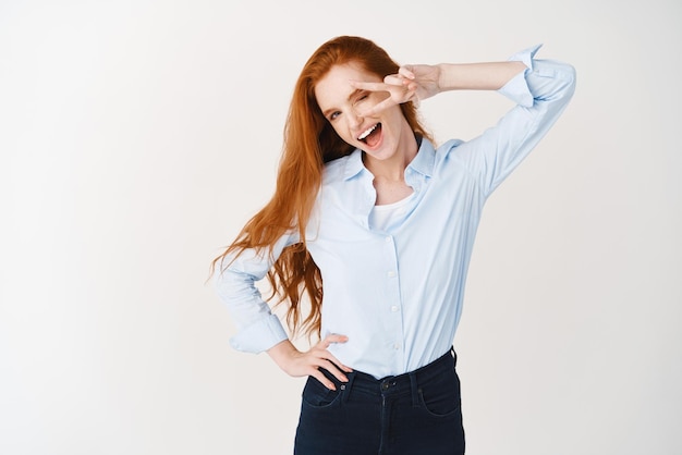 Image of young ginger girl in blue shirt showing peace sign isolated over white background