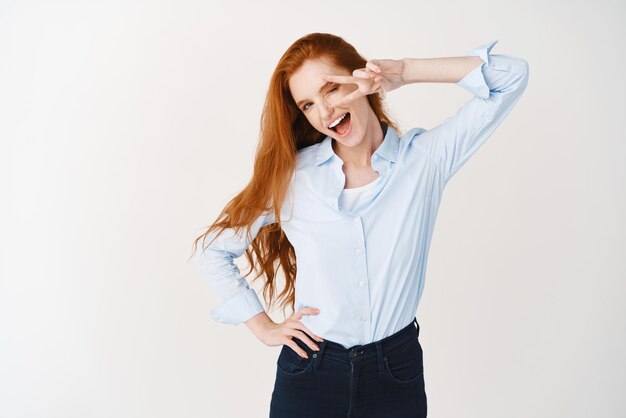 Image of young ginger girl in blue shirt showing peace sign isolated over white background