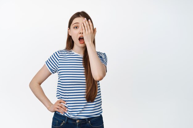 Image of young girl without makeup covers half of face, one side and looking surprised at camera, standing over white background