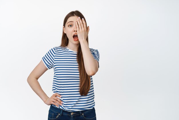 Image of young girl without makeup covers half of face one side and looking surprised at camera standing over white background