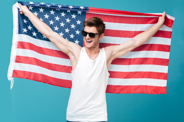 Image of young handsome man standing over blue isolated  holding USA flag. Looking aside.