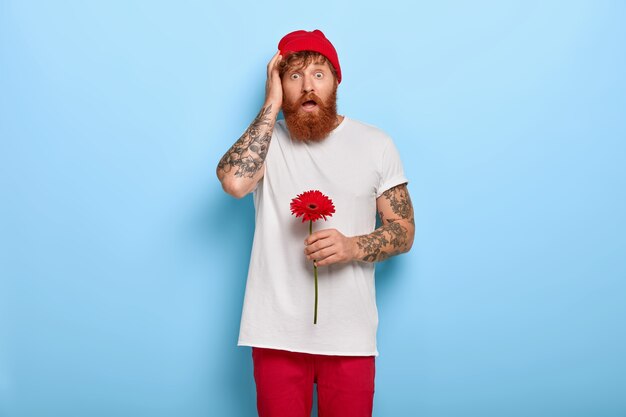 Image of young hipster holds red gerbera flower, comes on date, shocked to notice girlfriend with other man