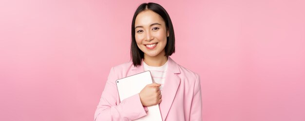 Image of young korean corporate woman ceo manager holding digital tablet smiling and looking professional wearing suit standing over pink background