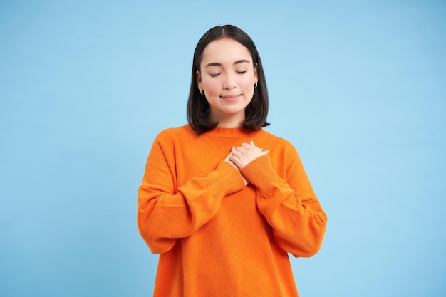 Image of young korean woman closes her eyes holds hands on hearts thinking of something with care an