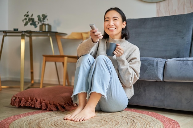Image of young korean woman drinks coffee holds remote from tv watching television at home resting i