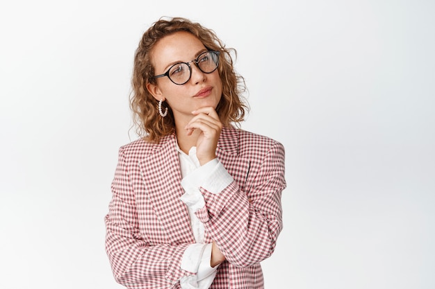 Image of young lady boss in glasses and suit thinking, makes decision, touches chin and looks thoughtful on white.