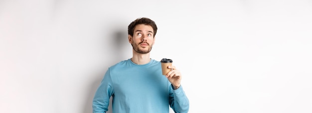 Free photo image of young man drinking takeaway coffee and looking up with thoughtful face tasting new flavour