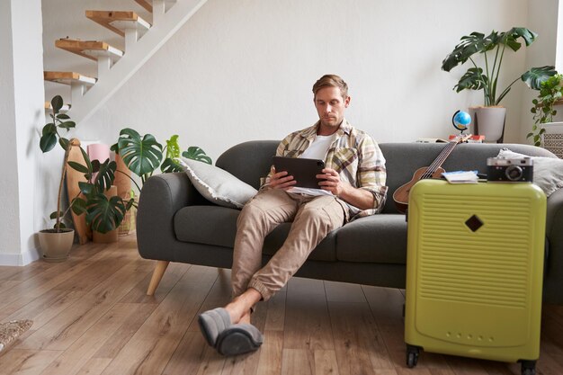 Free photo image of young man with tablet sitting in living room with suitcase reading studying the route