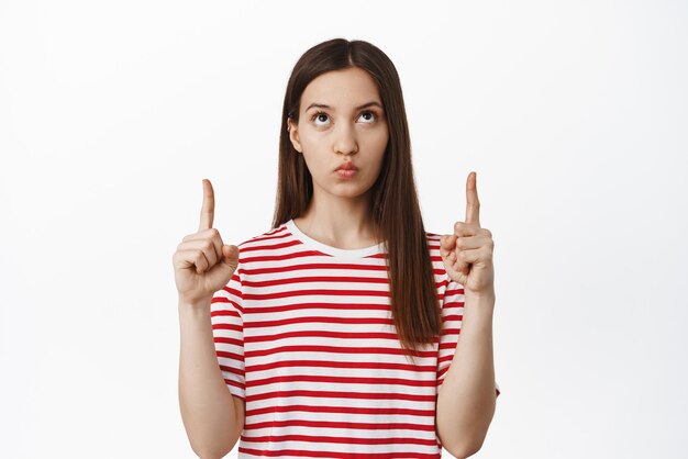 Изображение молодой угрюмой девушки, указывающей пальцем вверх и смотрящей на что-то с озадаченным задумчивым лицом, думающей о чем-то стоящем в красной футболке на белом фоне