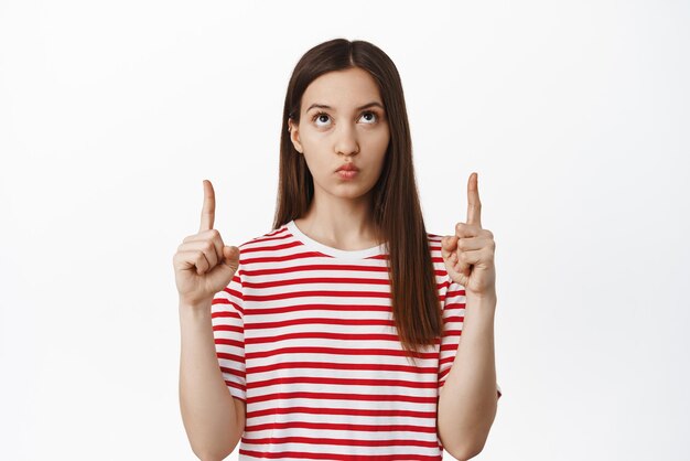 Image of young moody girl pointing fingers up looking at smth with puzzled thoughtful face thinking of something standing in red tshirt against white background