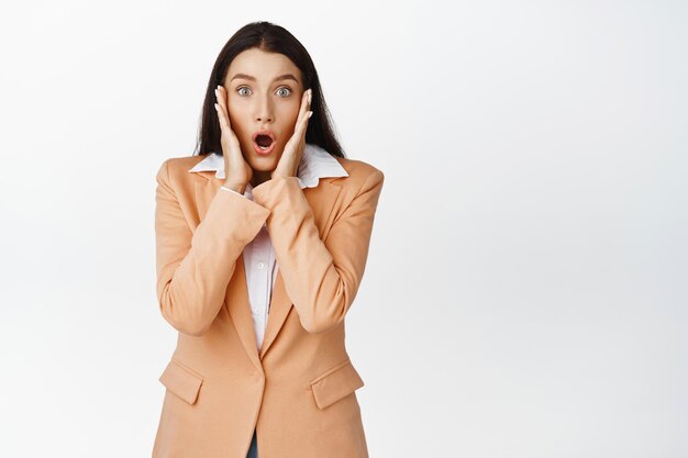 Image of young office woman looks surprised gasping excited and staring at camera hear shocking news in office standing over white background