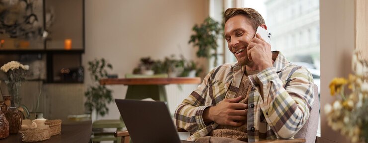 Free Photo image of young professional cafe visitor working sitting with laptop and talking on mobile phone to client a guy making an enquiry buying something online calling a friend
