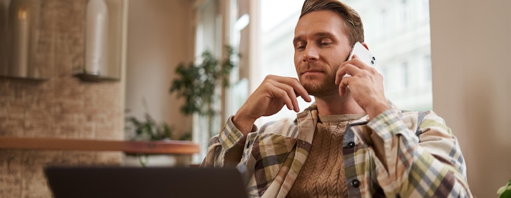 Free Photo image of young professional cafe visitor working sitting with laptop and talking on mobile phone to client a guy making an enquiry buying something online calling a friend