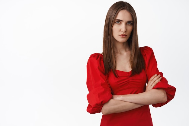Image of young serious and confident young in red trendy dress, cross arms on chest, looks bossy and self-assured, standing displeased against white background