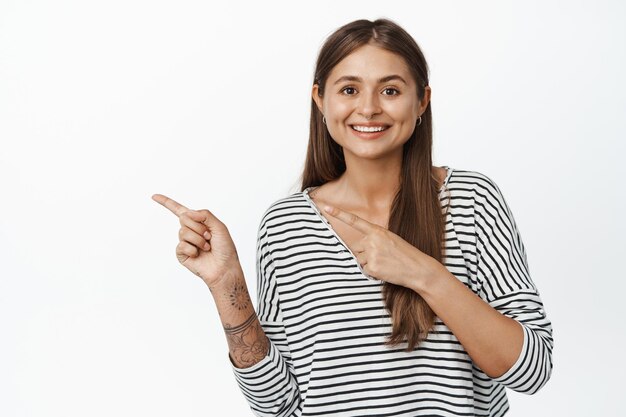 Image of young smiling woman 25s, pointing fingers left at logo, showing banner or advertisement copy space, standing against white background.