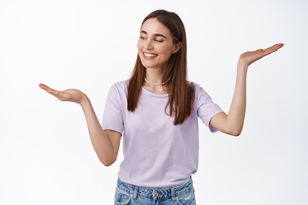 Image of young smiling woman shopping, holding in both hands, look at open palm as if making decision, choose what to buy, pick from two variants, white background