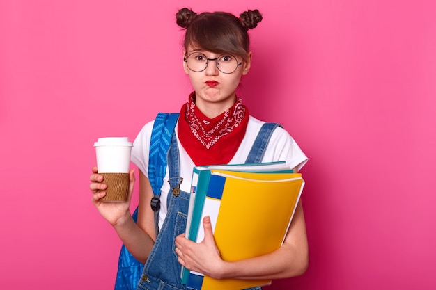 Image of young student holding clipboard and coffee cup while thinking about her report, feels disappointment, dislikes topic for her essay, being in bad mood, wearing fashionable clothes.