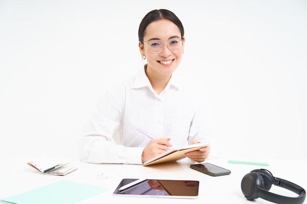 Free photo image of young tutor asian woman teacher sits in office writing working on report at workplace wears