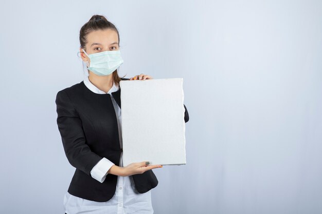 Image of young woman in face mask keeping blank canvas over white wall. High quality photo