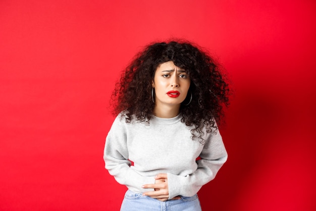 Immagine di una giovane donna che ha mal di stomaco, si piega per il dolore e si lamenta di crampi mestruali dolorosi, in piedi su sfondo rosso