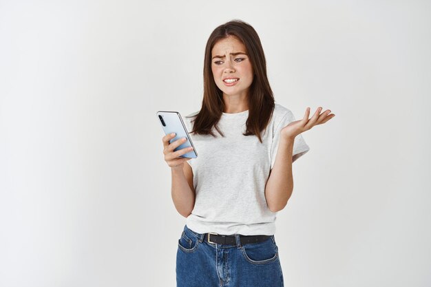 スマートフォンでぎこちなく不快に見える若い女性の画像、オンラインでクリンジメッセージを受信し、電話の画面を見つめ、肩をすくめる、白い壁